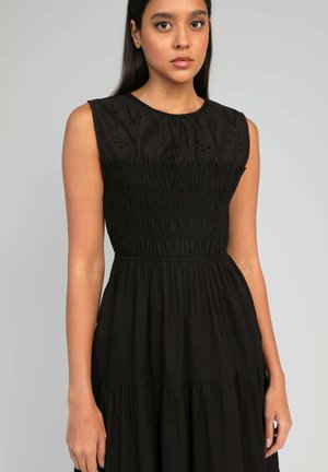 Jeune femme aux longs cheveux noirs portant une robe noire sans manches avec un corsage smocké et des détails en dentelle sur le haut, debout devant un fond uni.