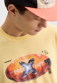 Žluté bavlněné tričko s grafickým potiskem v oranžových a černých akcentních barvách, zobrazující retro herní prvky a text značky.