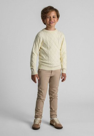 Pull en tricot crème avec un motif en losange, pantalon beige et baskets marron. La texture est lisse ; le pull présente des poignets et un ourlet côtelés.