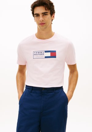 Tânăr purtând un tricou roz pal Tommy Hilfiger cu logo și pantaloni bleumarin, stând cu mâinile în buzunare pe fundal alb.