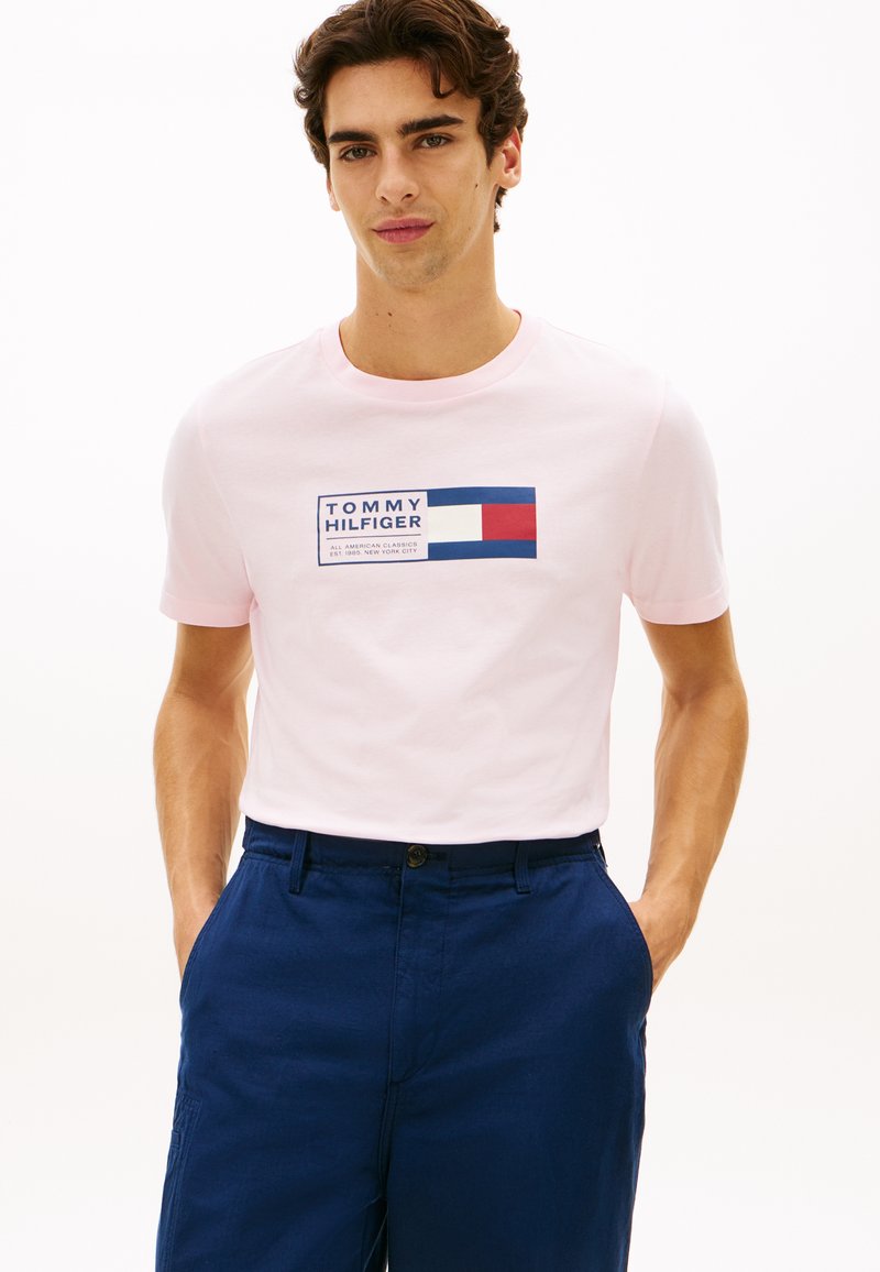 Νέος άνδρας με ανοιχτό ροζ μπλουζάκι Tommy Hilfiger με λογότυπο και σκούρο μπλε παντελόνι, στέκεται με τα χέρια στις τσέπες μπροστά σε λευκό φόντο.