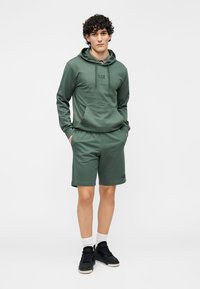 Zielona bawełniana bluza z kapturem z logo, zestawiona z pasującymi szortami. Model nosi czarne trampki i białe skarpetki do kostek. Prosty, casualowy design.