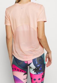 Licht perzikkleurig sport-T-shirt met mesh inzetstukken aan de achterkant. Korte mouwen en een afgeronde zoom. Gecombineerd met kleurrijke, patroonleggings.