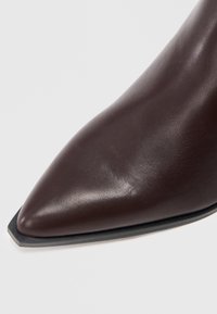 Hnědá kožená vysoká bota na podpatku s špičatou špičkou a elegantním designem. Má hladkou texturu a kontrastní černou podrážku.