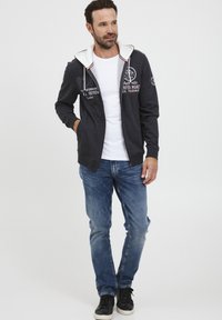 Tamno siva zip-up majica s bijelim unutarnjim oblogom, grafičkim dizajnom, u kombinaciji s plavim trapericama i crnim tenisicama. Opusti fit i ležeran stil.