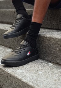 Sneakers in pelle nera con dettagli perforati, suola piatta e punta arrotondata. Presentano una piccola etichetta logo in rosso, bianco e blu sul lato.
