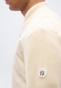 Άνδρας με εκρού πουλόβερ ζιβάγκο με πλεκτά μανίκια και μικρό στρογγυλό μεταλλικό λογότυπο στο επάνω μέρος του μπράτσου, πάνω σε απλή φόντο.