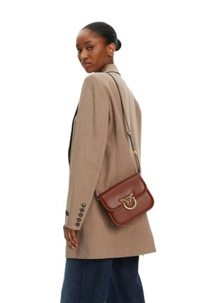 Donna che indossa un blazer beige e jeans scuri, con una borsa a tracolla in pelle marrone con chiusura dorata a forma di farfalla.