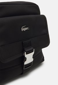 Borsa a tracolla nera in tessuto con chiusura a patta, dotata di un gancio in metallo argentato e un logo a forma di coccodrillo impresso sul davanti. Materiale texturizzato.