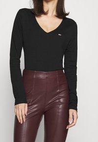 Svart långärmad topp med V-ringning och en liten logopatch. Kombinerad med höga burgundyröda byxor i konstläder.