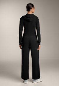 Fekete overál, amelynek fitted felső része és bő szára van. Kapucnival, hosszú ujjakkal és sima anyaggal rendelkezik; fehér tornacipővel viselve.