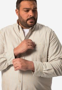 Une chemise à manches longues avec un tissu beige clair à fines rayures bleues, une poche avant et des détails de boutons sur les poignets.