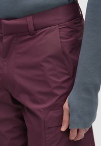 Pogled od blizu na osebo, ki nosi hruškaste hlače s žepom in temno sivo dolg rokav s odprtinami za prste.