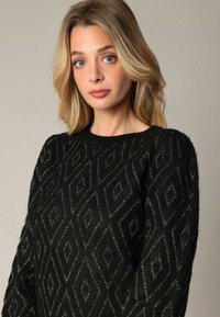 Femme blonde aux yeux bleus portant un pull noir avec un motif en losange texturé, posant devant un fond uni.