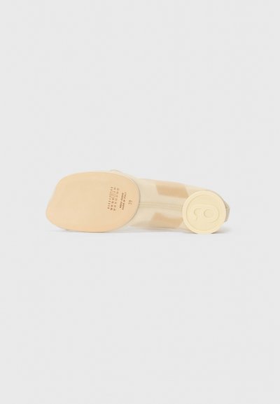 Beige shoe with a soft, smooth upper material, rounded toe, and a circular logo detail on the sole. The bottom is flat with a minimal tread.