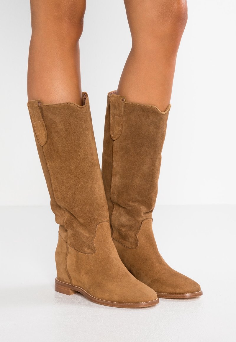 Bottes compensées en daim marron montant jusqu'aux genoux, portées sur des jambes nues, avec des détails à languette en haut et des talons bas en bois.