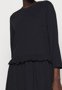 Svart långärmad klänning i mjukt tyg, med volangdetalj i midjan och rund halsringning för en avslappnad passform.