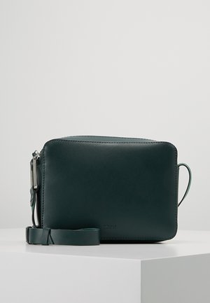 Sac bandoulière rectangulaire en cuir vert foncé avec sangle réglable et fermeture éclair, posé sur un piédestal blanc sur fond uni.