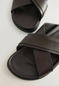 Sandales en cuir marron avec des sangles entrecroisées et une semelle plate. Fini texturé sur les sangles ; surface de la semelle lisse ; design minimaliste.