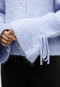 Helllavendelfarbener, gestrickter Pullover mit breiten, strukturierten Ärmeln und einem Zugband-Detail, das eine weiche, flauschige Oberfläche und einen einzigen Knopfverschluss aufweist.
