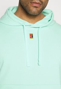 Sweat à capuche vert menthe en tissu doux, avec une poche avant, une capuche avec cordon de serrage et un patch logo multicolore sur la poitrine.