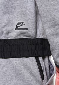 Šedá mikina se černým elastickým pasem, obsahuje nášivku s logem Nike; má zipovou kapsu a strukturovanou tkaninu.