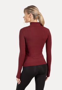 Borgå-röd träningsöverdel med lång ärm, hög krage och tajt passform. Mjuk yta med sömdetaljer, kombinerad med svarta leggings.