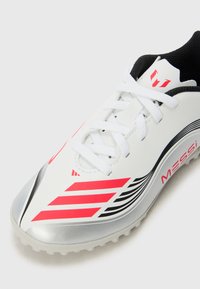 Crampons de soccer blancs avec des accents noirs, des rayures roses et une semelle argentée. Présente un design texturé et un système de laçage pour un ajustement sécurisé.