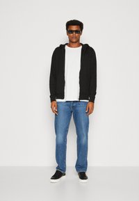 Schwarze Kapuzenjacke über einem weißen T-Shirt, kombiniert mit blauen Jeans und schwarzen Sneakers. Das Model trägt eine dunkle Sonnenbrille.