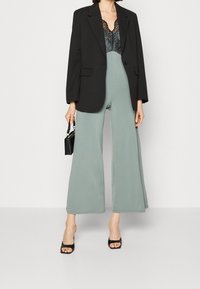 Černý blazer přes krajkový body, spárovaný se zeleným širokým overalem. Modelka drží malou černou kabelku a má na sobě černé sandály na podpatku.