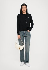 Cardigan nero con bottoni, indossato sopra una maglietta nera. Abbinato a jeans blu chiaro e sneakers bianche, tiene in mano una borsa bianca a forma curva.