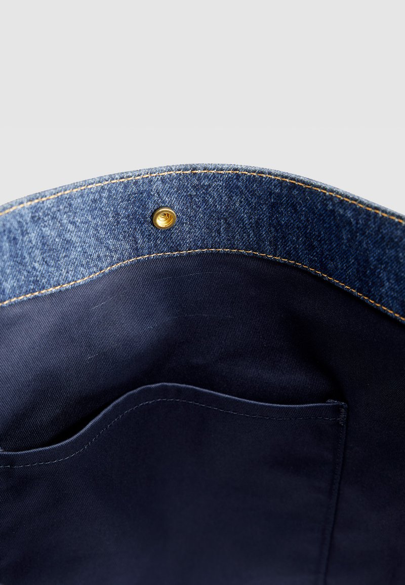 Innenseite einer Jeans-Tasche mit dunkelblauem Stofffutter, einer Innentasche, kontrastierenden gelben Nähten und einem Messing-Druckknopfverschluss.
