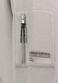 Gray sweatshirt with a zippered pocket featuring black cord accents. A label reads "URBAN SURVIVAL ORIGINAL COLLECTION SUB. LEVEL."