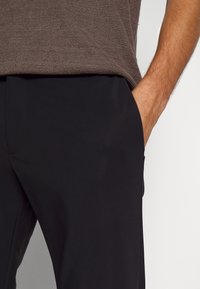 Homme portant une chemise texturée marron foncé et un pantalon noir, la main droite dans la poche, sur un fond blanc uni.