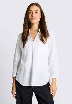 Kobieta z długimi brązowymi włosami, ubrana w białą koszulę na guziki i ciemne spodnie, stojąca na jasnym, jednolitym tle.