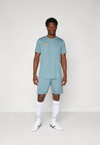Camisola e calções desportivos azul-claro feitos de tecido liso, com um logótipo dourado na camisola. Meias brancas até ao joelho e sapatilhas desportivas.