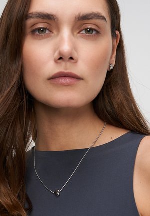 Silver necklace with a smooth round pendant, worn over a navy sleeveless top. Model has long brown hair and minimal makeup.