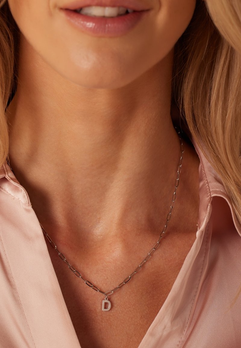Zilveren ketting driehoekjes met verlengde schakels en een kleine hanger met de letter "D." Draagt over een lichtroze stof.