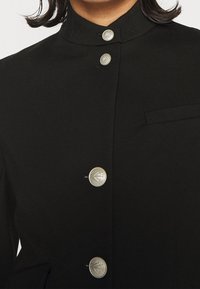 Prenda negra con un cuello alto, botones plateados y un único bolsillo en el pecho, con líneas limpias y estructuradas y una textura suave.