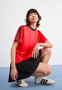 Raudona sportinė maikutė su juodais akcentais, juoda klostuota sijonas ir kreminės spalvos sportiniai bateliai su juodais dryžiais. Lygus paviršius ir laisvas siluetas.