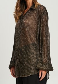 Blusa trasparente a maniche lunghe in un tessuto scuro e iridescente con strisce verticali, dotata di frontale con bottoni e colletto arrotondato.
