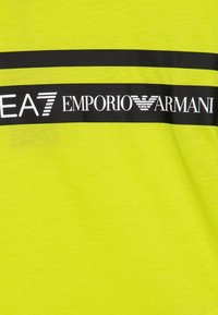 Neon žluté tričko s černým horizontálním pruhem, na kterém je bílý nápis "EA7 EMPORIO ARMANI", hladká textura a kulatý výstřih.