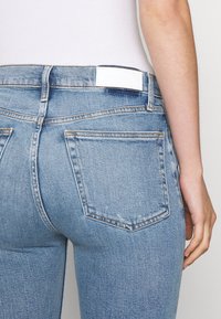 Jeans de mezclilla azul claro con un diseño ajustado. Las características incluyen una etiqueta blanca en la cintura, dos bolsillos traseros y un desgaste sutil.