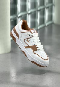 Λευκό και καφέ αθλητικό παπούτσι Lacoste με κορδόνια, λογότυπο και τρύπες, αιωρούμενο πάνω από ένα γυαλισμένο σκυρόδεμα κοντά σε γυάλινους τοίχους.