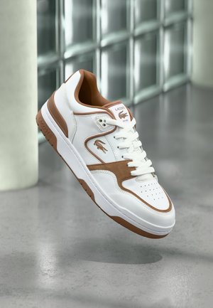 Sneakers Lacoste bianche e marroni con lacci, logo e perforazioni, sospese sopra un pavimento in cemento lucido vicino a blocchi di vetro.