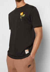 Svart t-shirt med kort ärm och orange Nike-logotyp, samt en grön bladdesign. Mjukt bomullsmaterial med en liten etikett på sid söm.