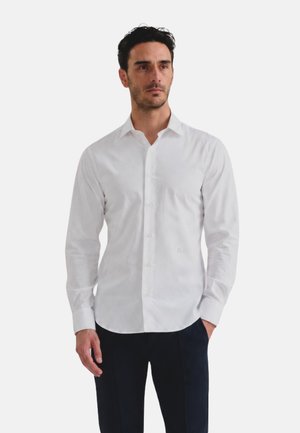 Uomo che indossa una camicia bianca a maniche lunghe con bottoni e pantaloni scuri, in piedi con una mano in tasca, sfondo neutro.