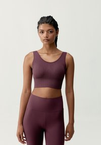 Mujer con el cabello trenzado, vistiendo un top corta de canalé en color ciruela y leggings de cintura alta a juego, de pie contra un fondo claro y liso.