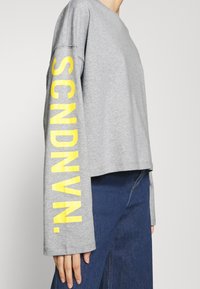Gray long-sleeve shirt with a cropped design, featuring yellow text "SCNDVN." printed vertically on the left sleeve. Soft fabric texture.