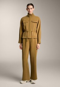 Compleu sport khaki cu guler înalt, buzunare frontale și talie cu șnur. Textură netedă și croială lejeră; asortat cu adidași de culoare deschisă.